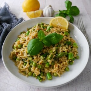 A white bowl of lemon orzo with asparagus and garden peas, garnished with fresh basil and served with a wedge of lemon.