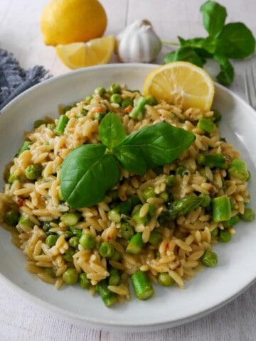 A white bowl of lemon orzo with asparagus and garden peas, garnished with fresh basil and served with a wedge of lemon.