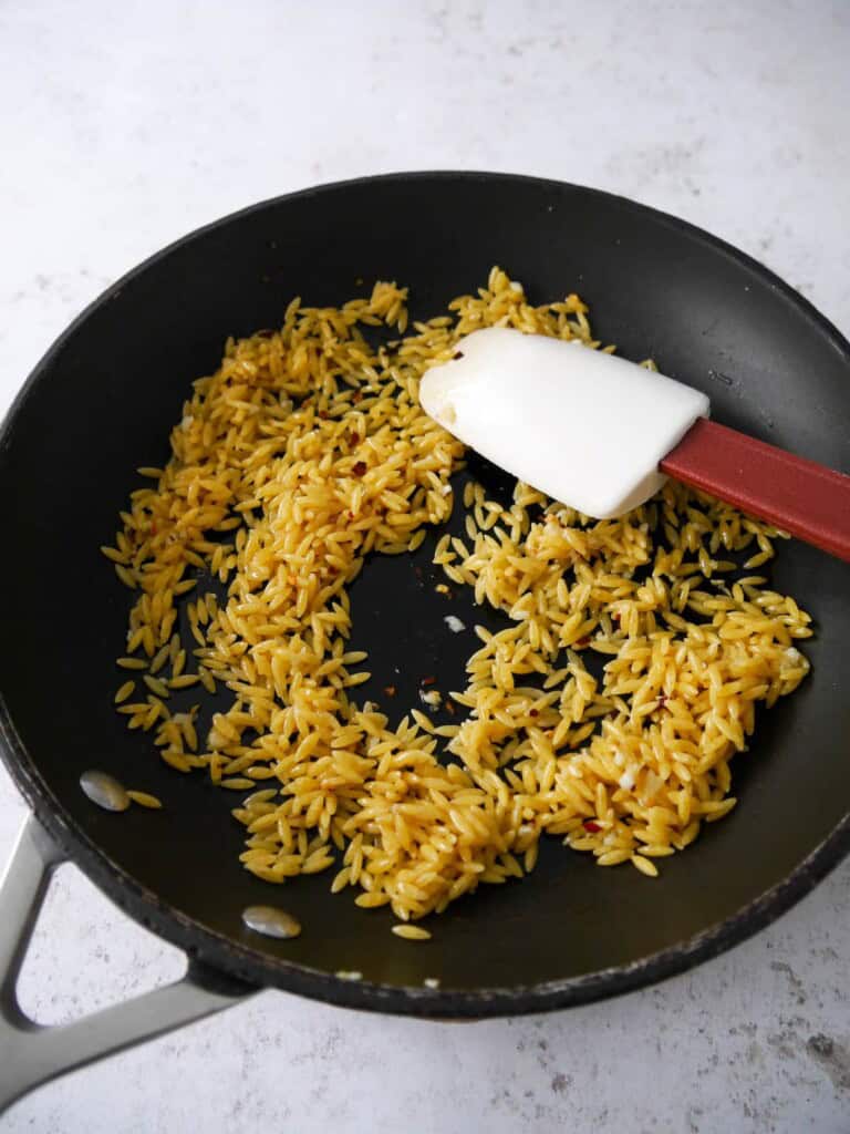 A non stick frying pan of olive oil, orzo pasta, garlic and chilli flakes.