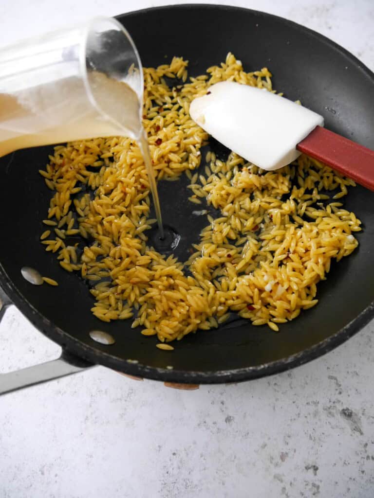 A non stick frying pan with orzo pasta, sauteed garlic and chilli flakes with vegetable stock being added.