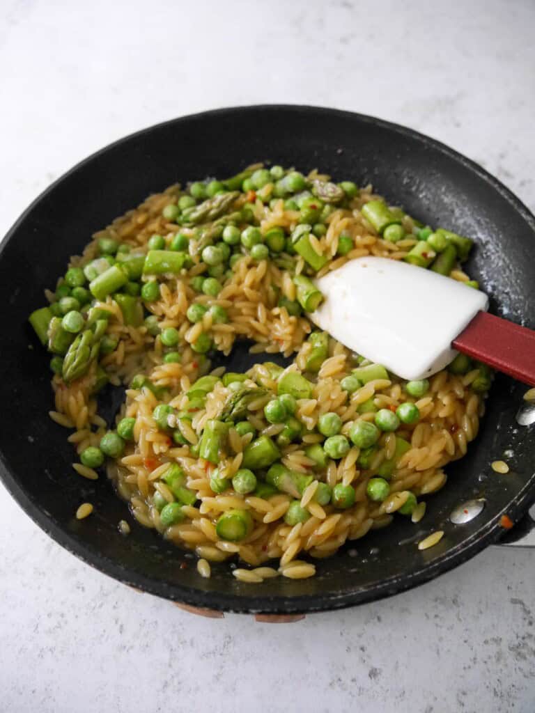 A non stick frying pan with cooked orzo pasta, asparagus and garden peas.