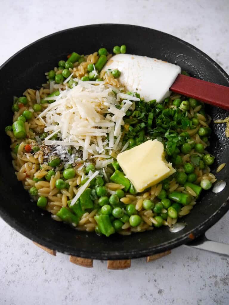 A non stick frying pan with cooked orzo, asparagus and peas with added chopped basil, parmesan cheese, lemon zest and juice, butter and seasoning.