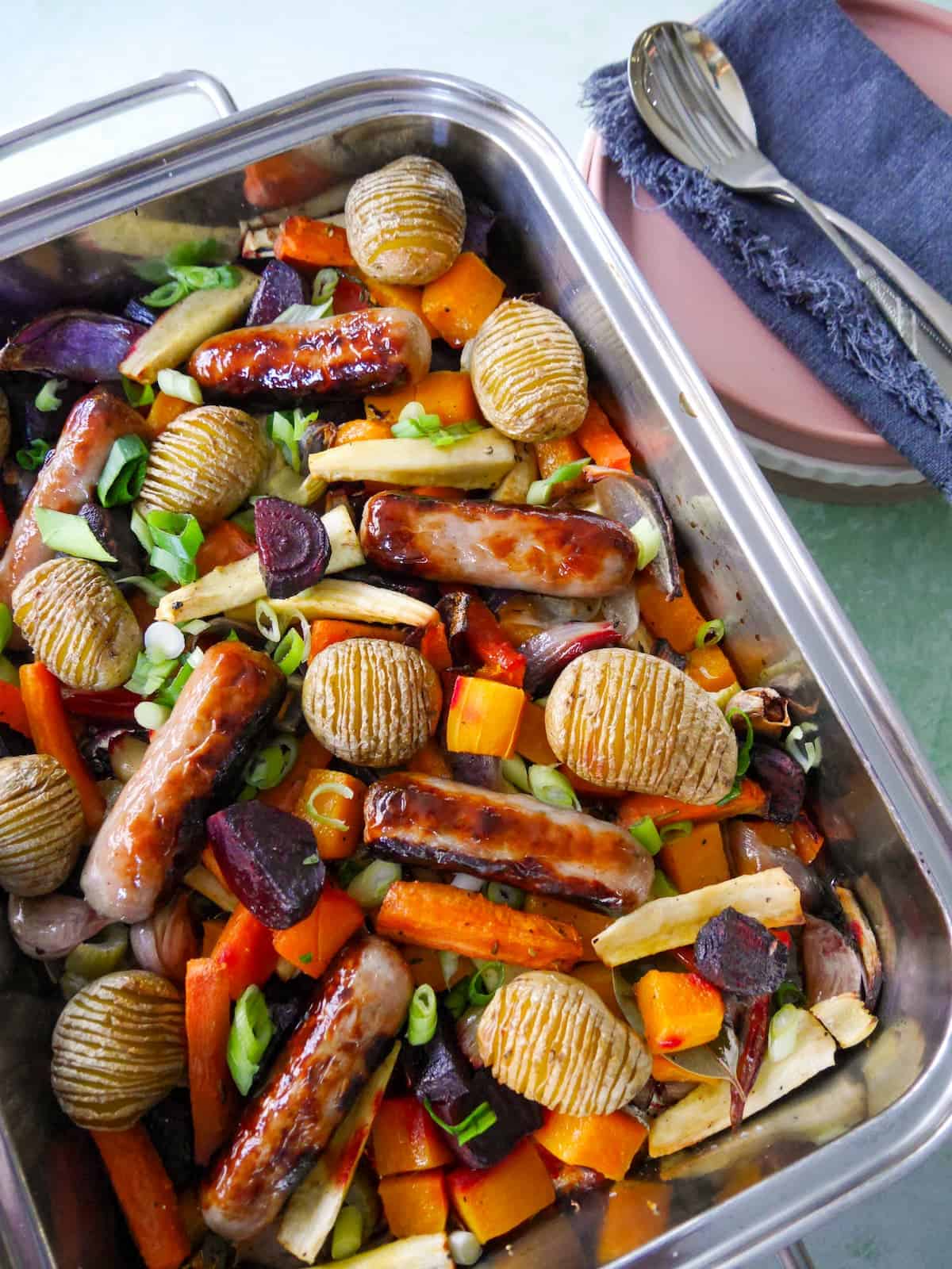 A large high sided baking tray filled with roasted root vegetables, hasselback potatoes and pork sausages.