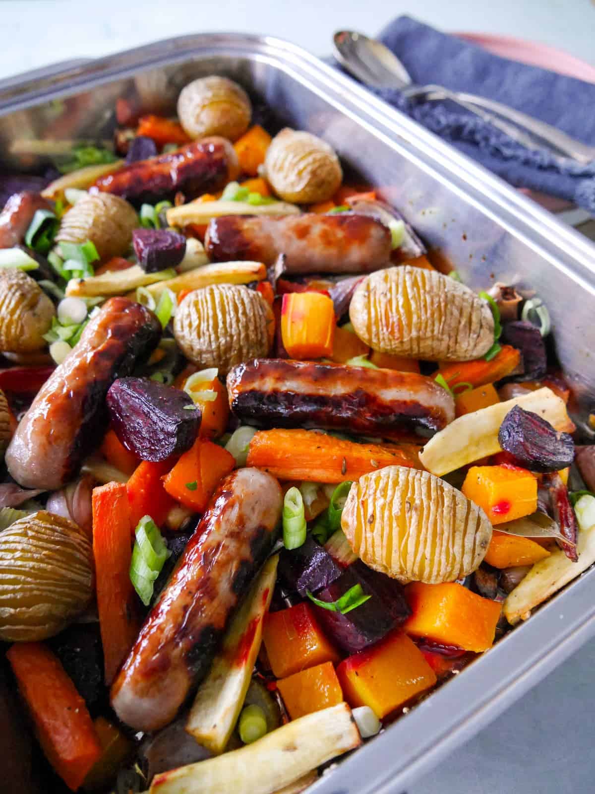 A large high sided baking tray filled with roasted root vegetables, hasselback potatoes and pork sausages.