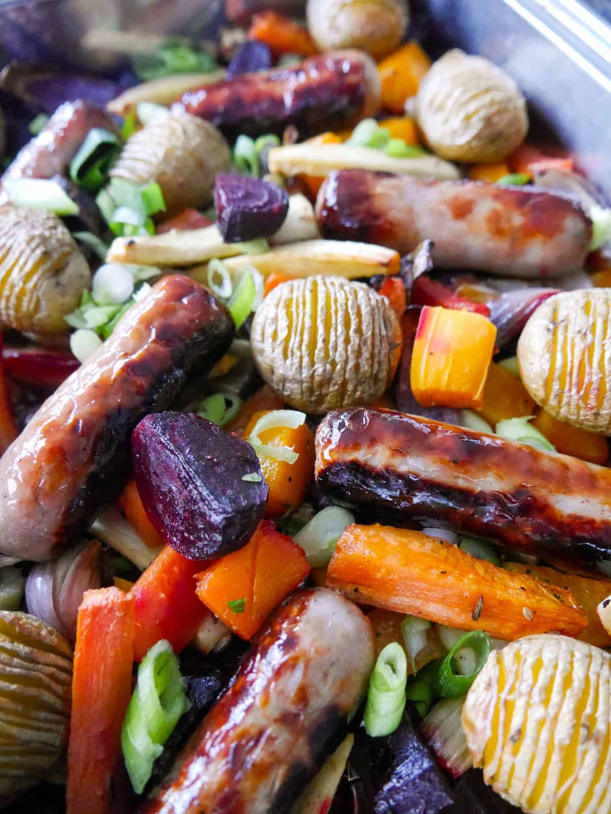 Close up image of roasted root vegetables, hasselback potatoes and pork sausages.