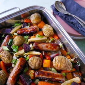 A large high sided baking tray filled with roasted root vegetables, hasselback potatoes and pork sausages.