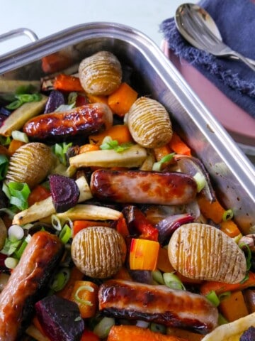 A large high sided baking tray filled with roasted root vegetables, hasselback potatoes and pork sausages.