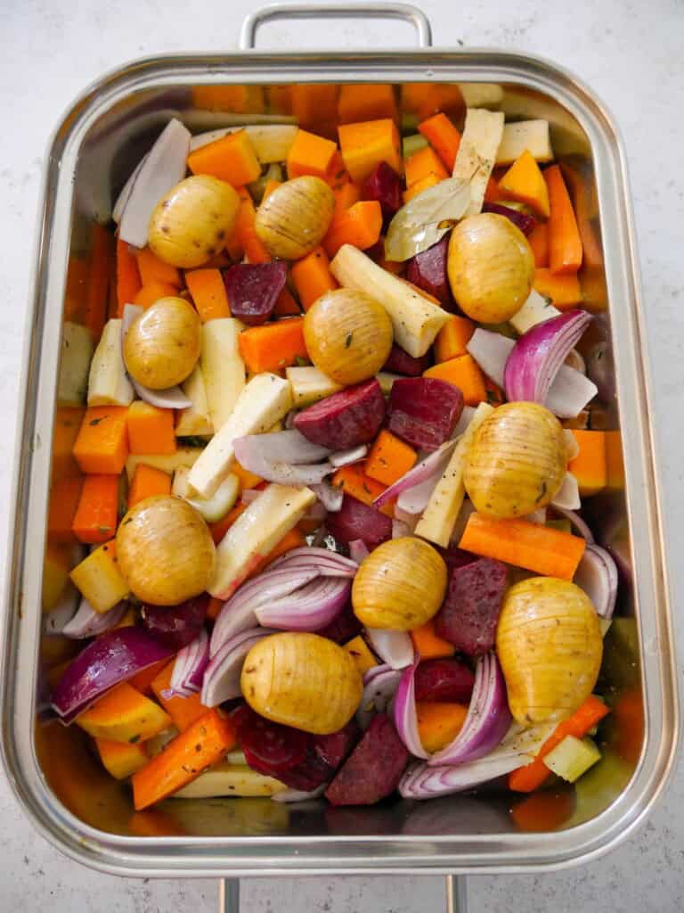 A large high sided baking tray filled with a variety of chopped root vegetables and hasselback baby potatoes.
