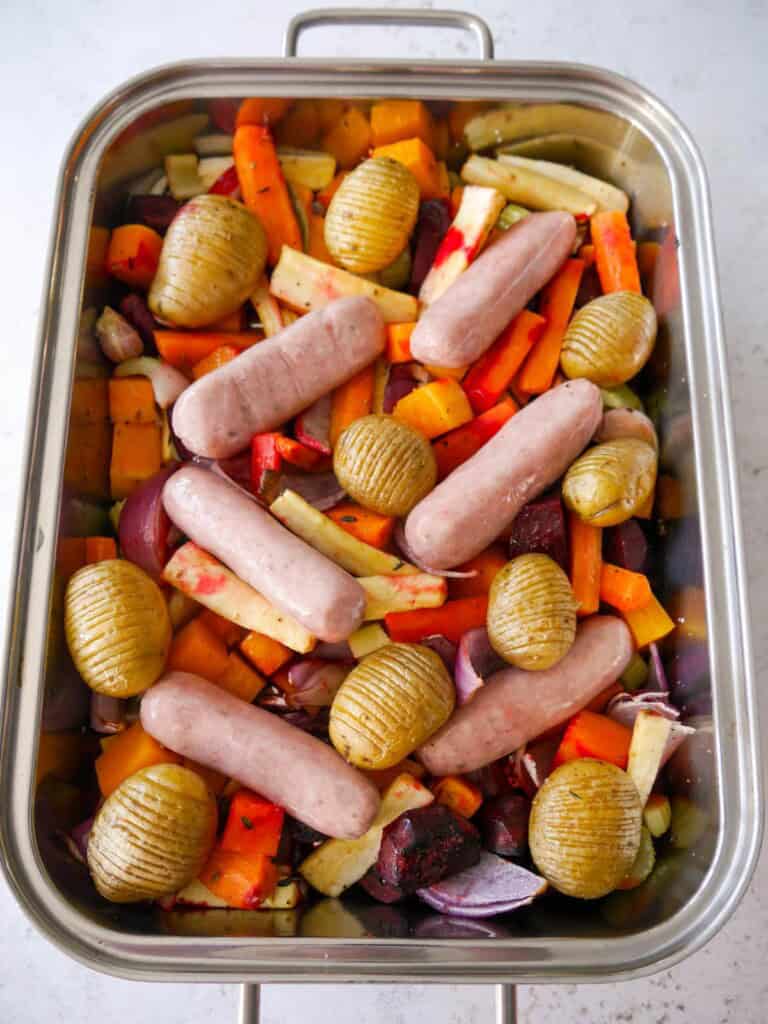 A large high sided baking tray filled with part roasted vegetables topped with raw pork sausages.