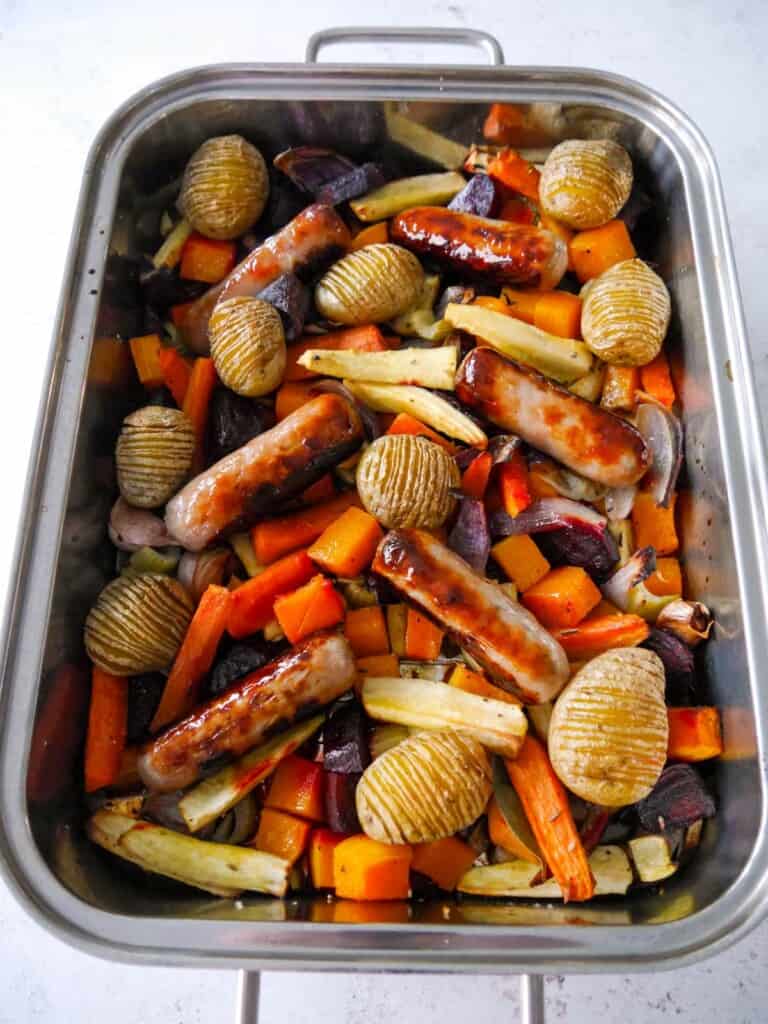 A large high sided baking tray filled with roasted root vegetables, hasselback potatoes and pork sausages.
