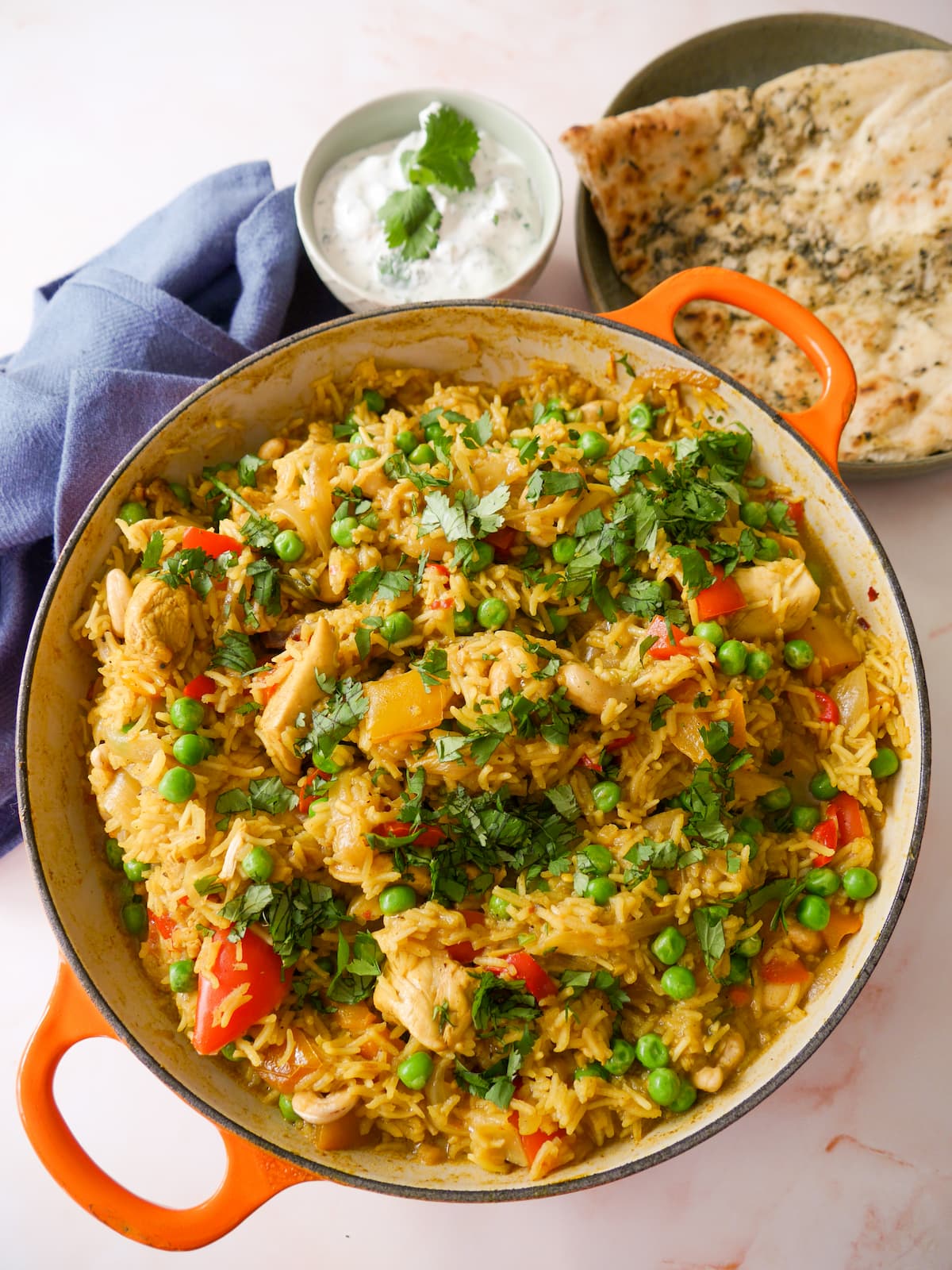 A large skillet of curried chicken and rice with added vegetables and cashew nuts, and garnished with coriander leaf served with garlic naan and yogurt raita.