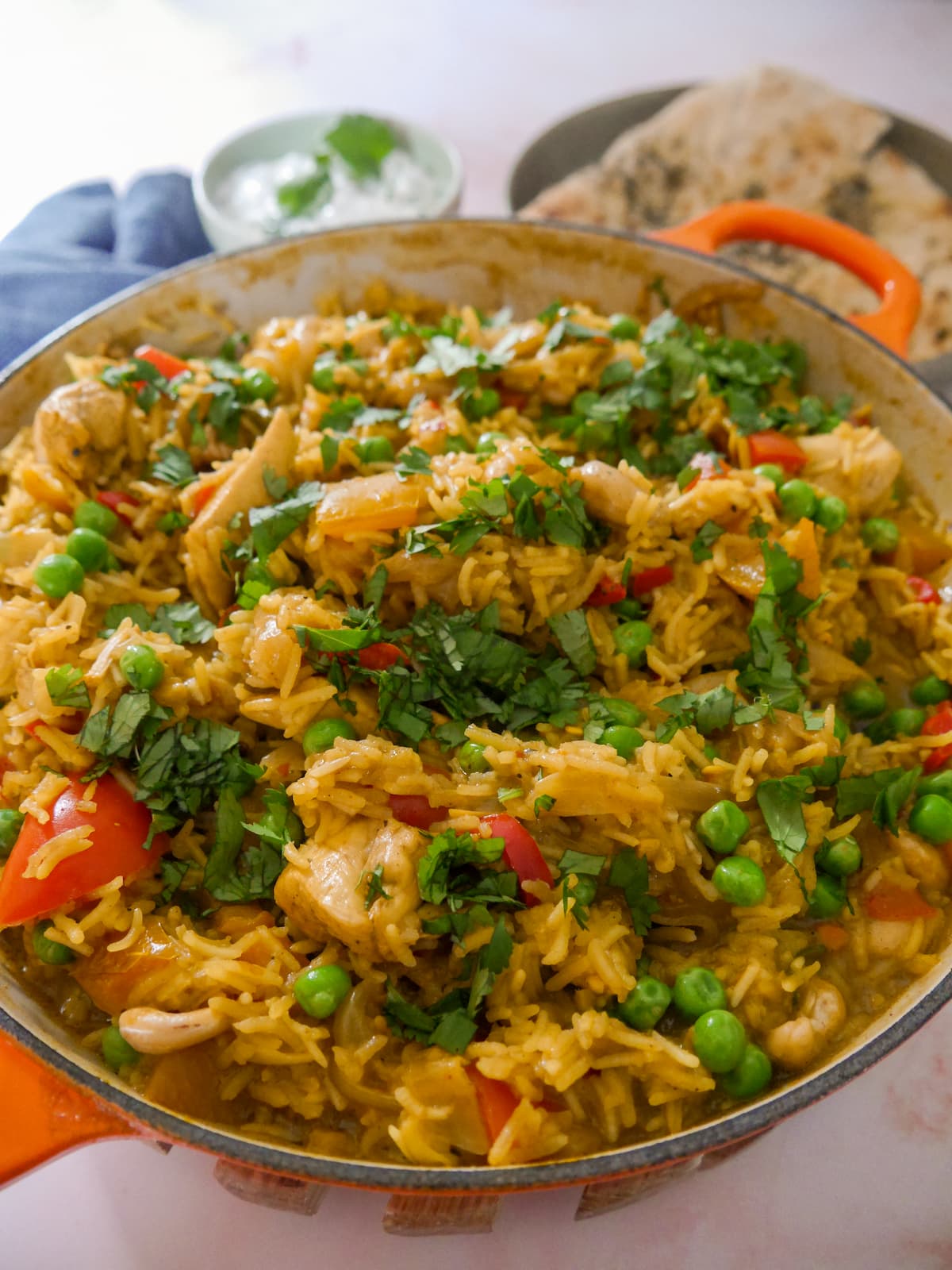 A large skillet of curried chicken and rice with added vegetables and cashew nuts, and garnished with coriander leaf served with garlic naan and yogurt raita.