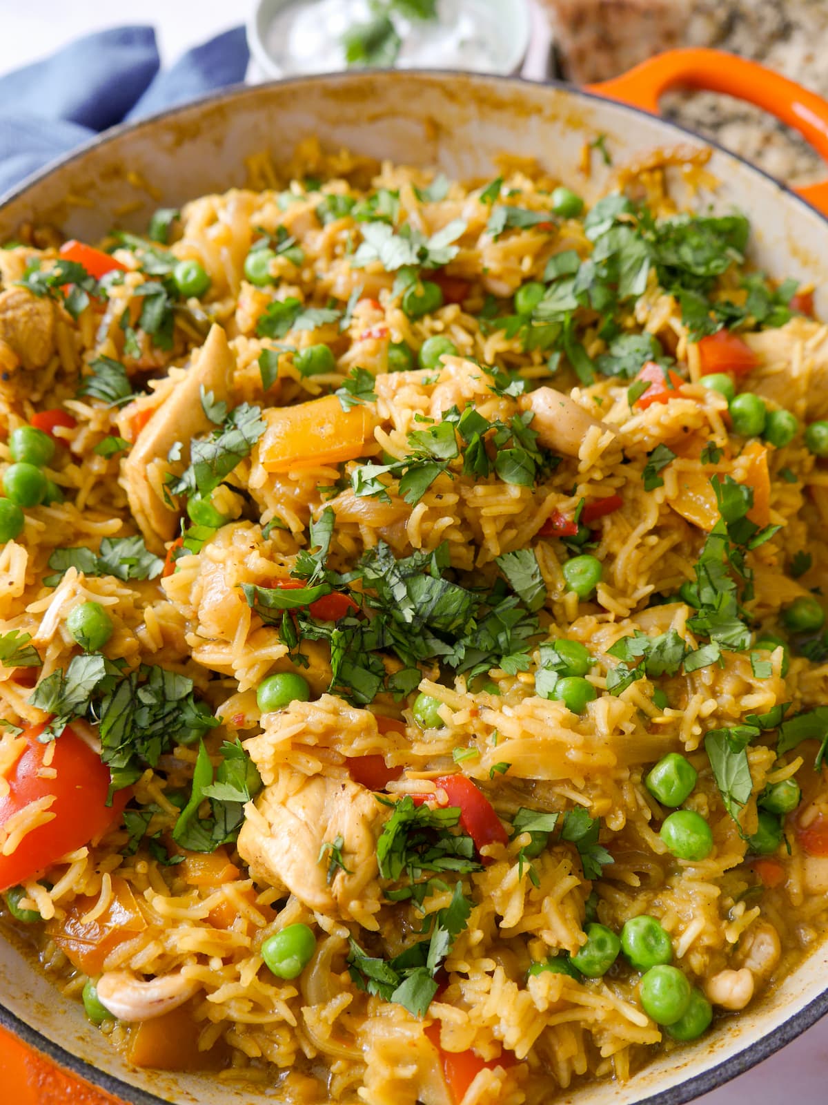 Close up image of a large skillet of curried chicken and rice with added vegetables and cashew nuts, and garnished with coriander leaf served with garlic naan and yogurt raita.