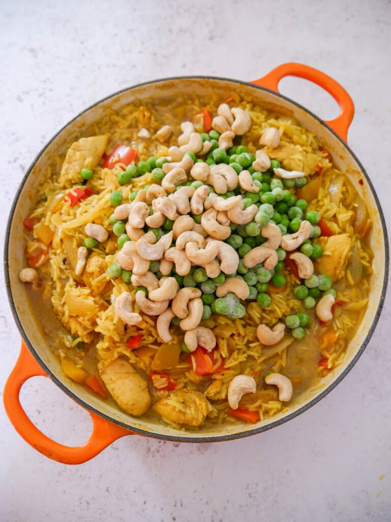 A large skillet with sauteed curried chicken and vegetables, cooked with rice and with garden peas and cashew nuts being added.