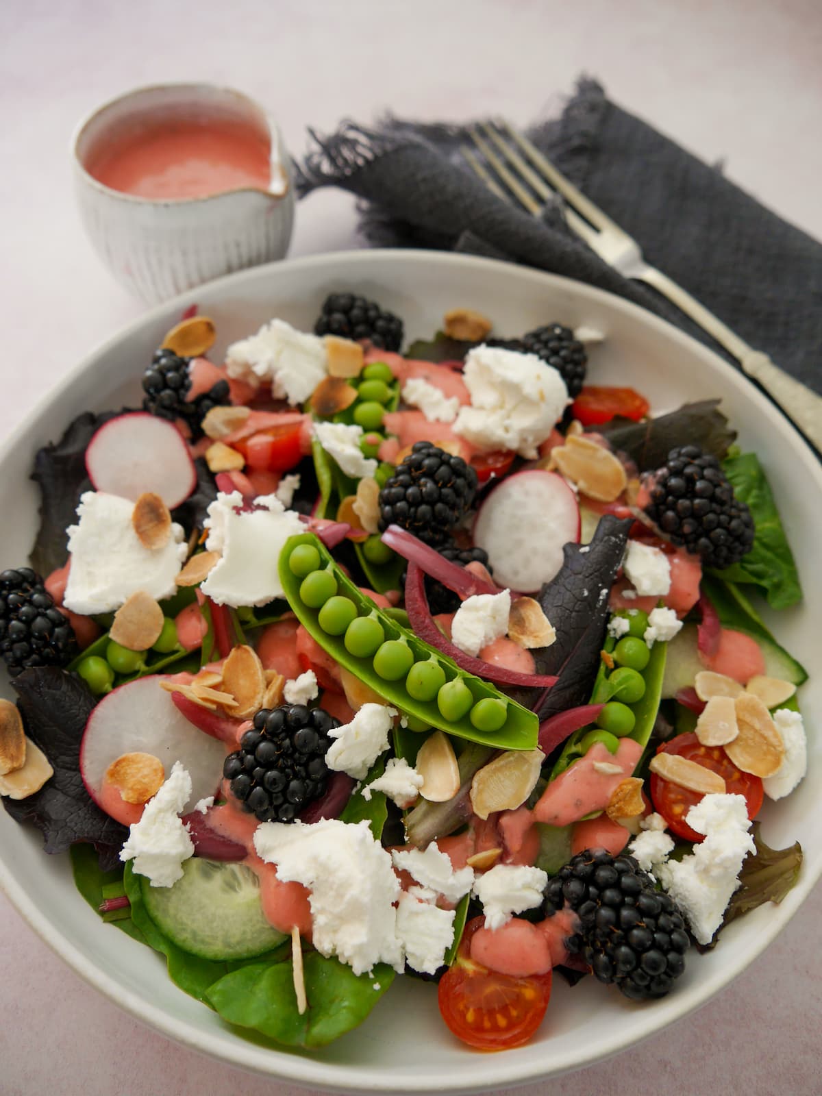 A bowl of mixed salad leaves, radish, tomato, cucumber, sugar snap peas, pickled red onions, with goat's cheese and blackberries served with a small jug of raspberry vinaigrette.