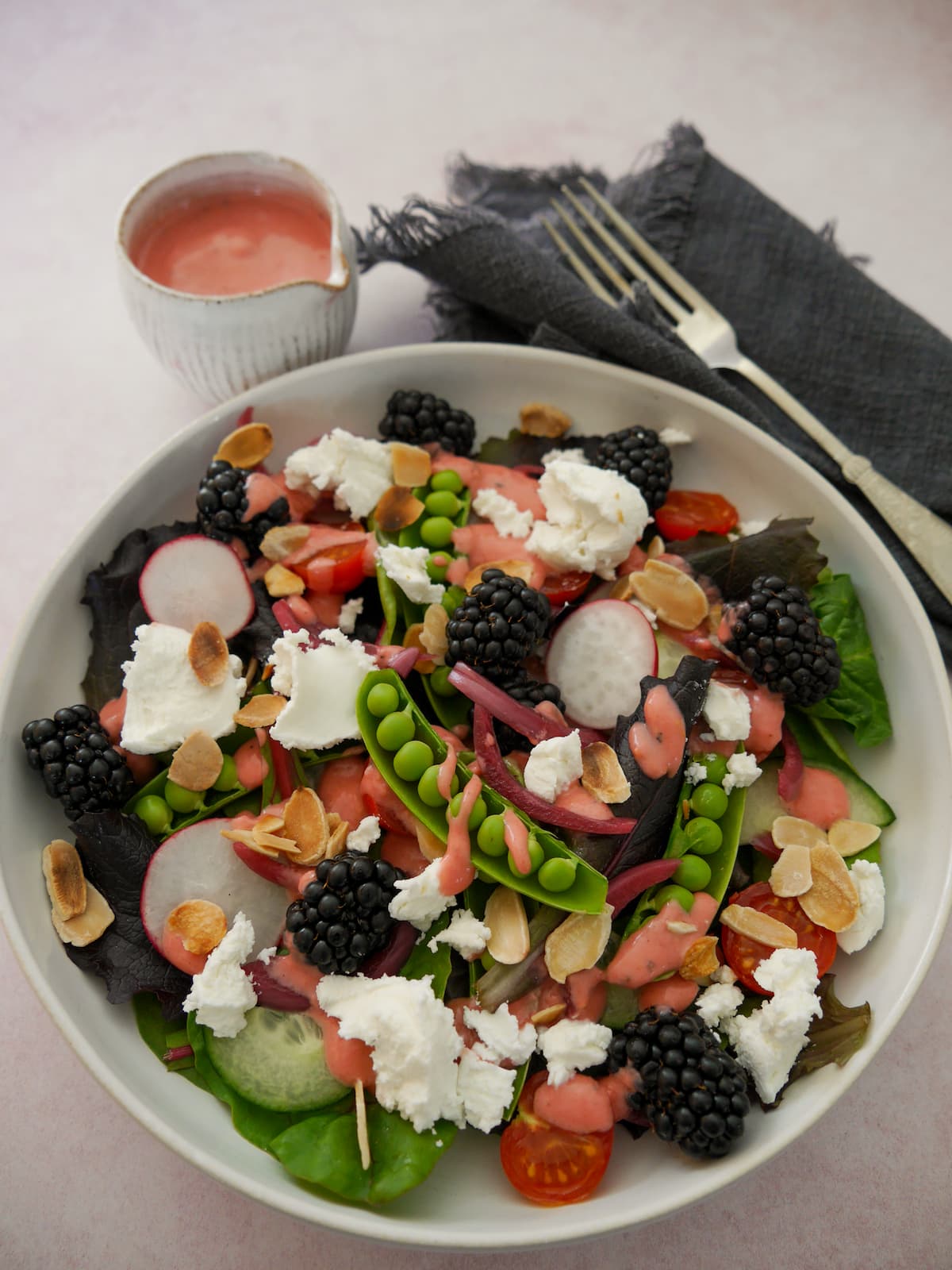 A bowl of mixed salad leaves, radish, tomato, cucumber, sugar snap peas, pickled red onions, with goat's cheese and blackberries served with a small jug of raspberry vinaigrette.