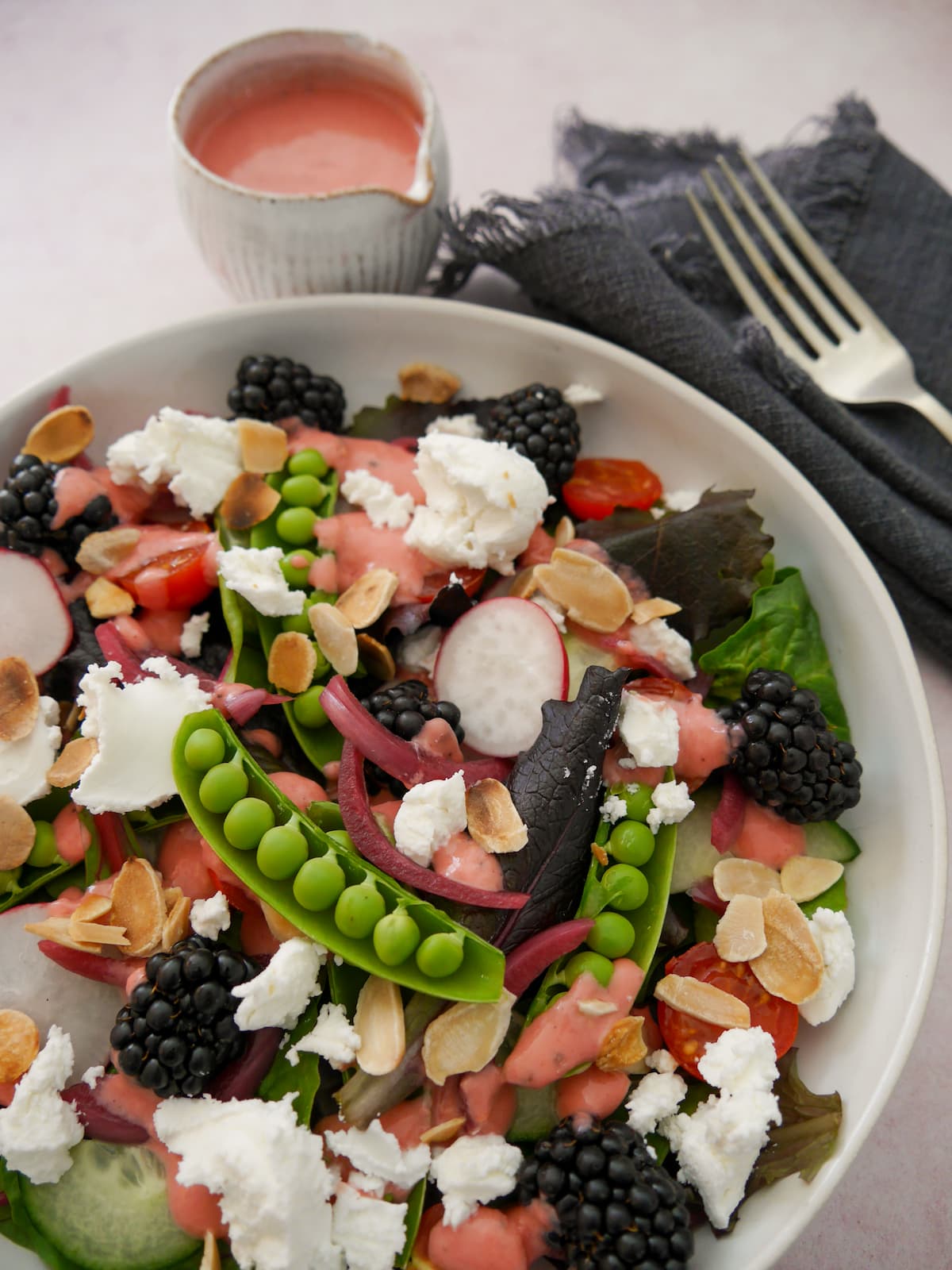 A bowl of mixed salad leaves, radish, tomato, cucumber, sugar snap peas, pickled red onions, with goat's cheese and blackberries served with a small jug of raspberry vinaigrette.