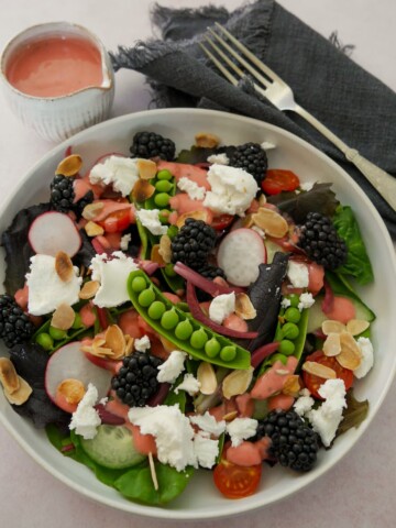 A bowl of mixed salad leaves, radish, tomato, cucumber, sugar snap peas, pickled red onions, with goat's cheese and blackberries served with a small jug of raspberry vinaigrette.