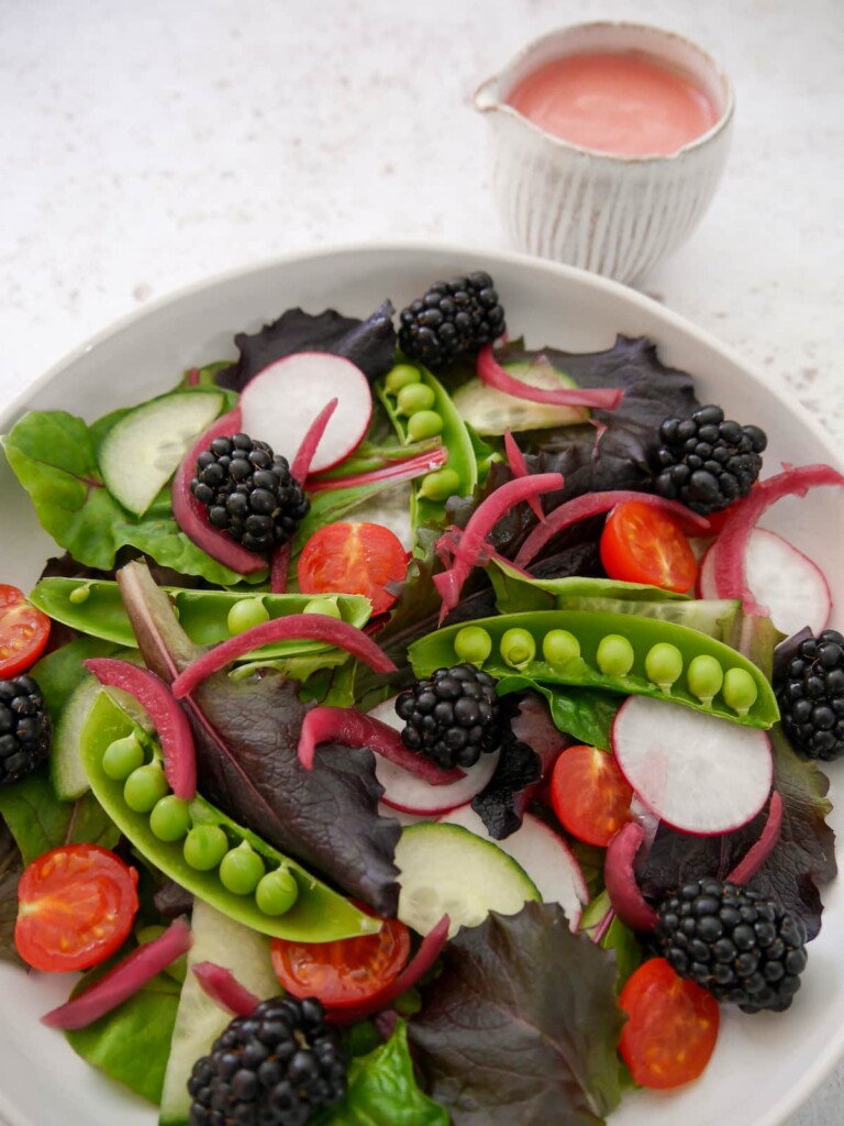 A bowl of mixed salad leaves, radish, tomato, cucumber, sugar snap peas, pickled red onions and blackberries, with a small jug of raspberry vinaigrette served alongside.