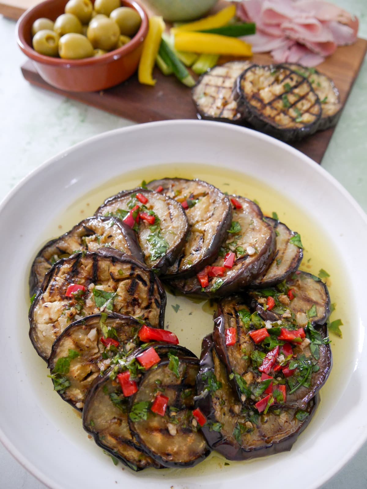 A bowl filled with Italian marinated aubergine slices, with a board of cold cuts and olives set alongside.
