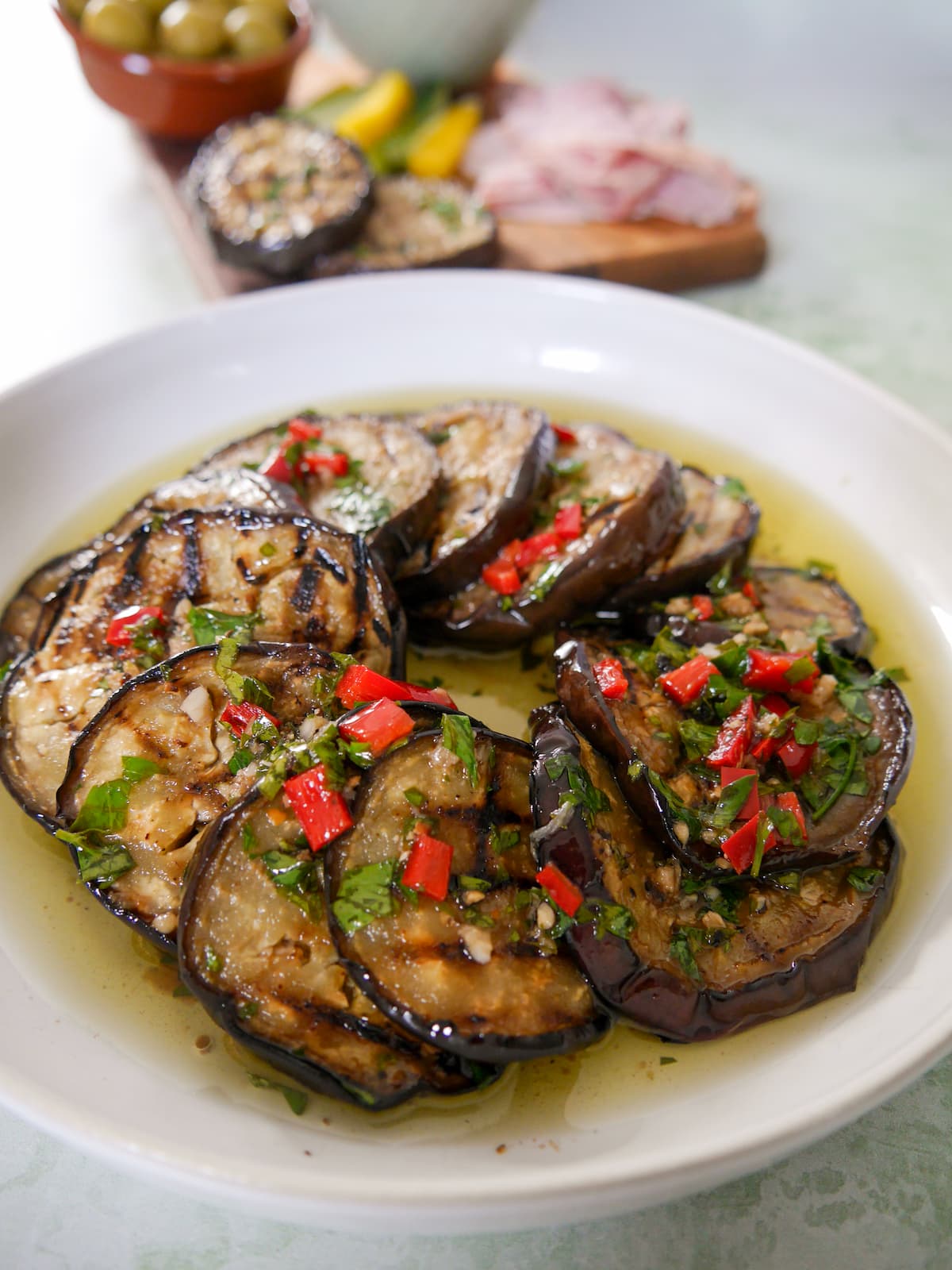 A bowl filled with Italian marinated aubergine slices, with a board of cold cuts and olives set alongside.