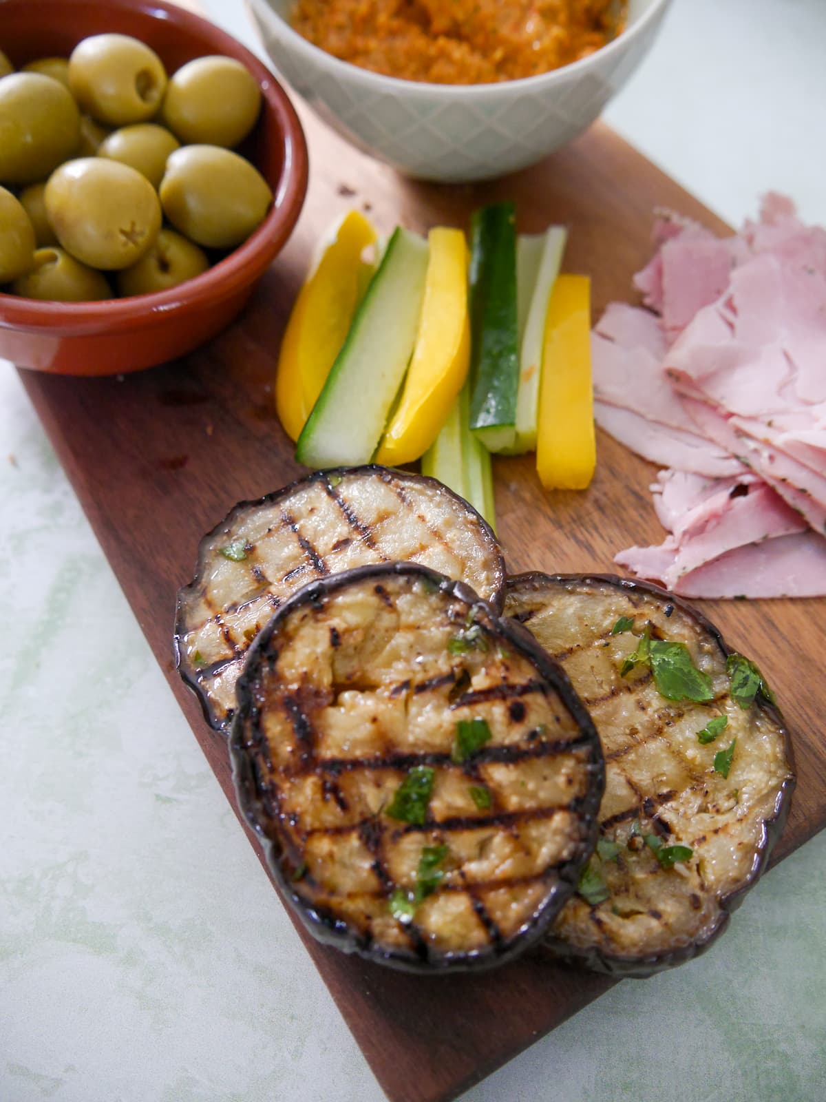 A wooden board topped with slices of Italian marinated aubergine, a bowl of green olives, a bowl of red pepper tapanade, slices of Italian ham and some batons of salad vegetables.