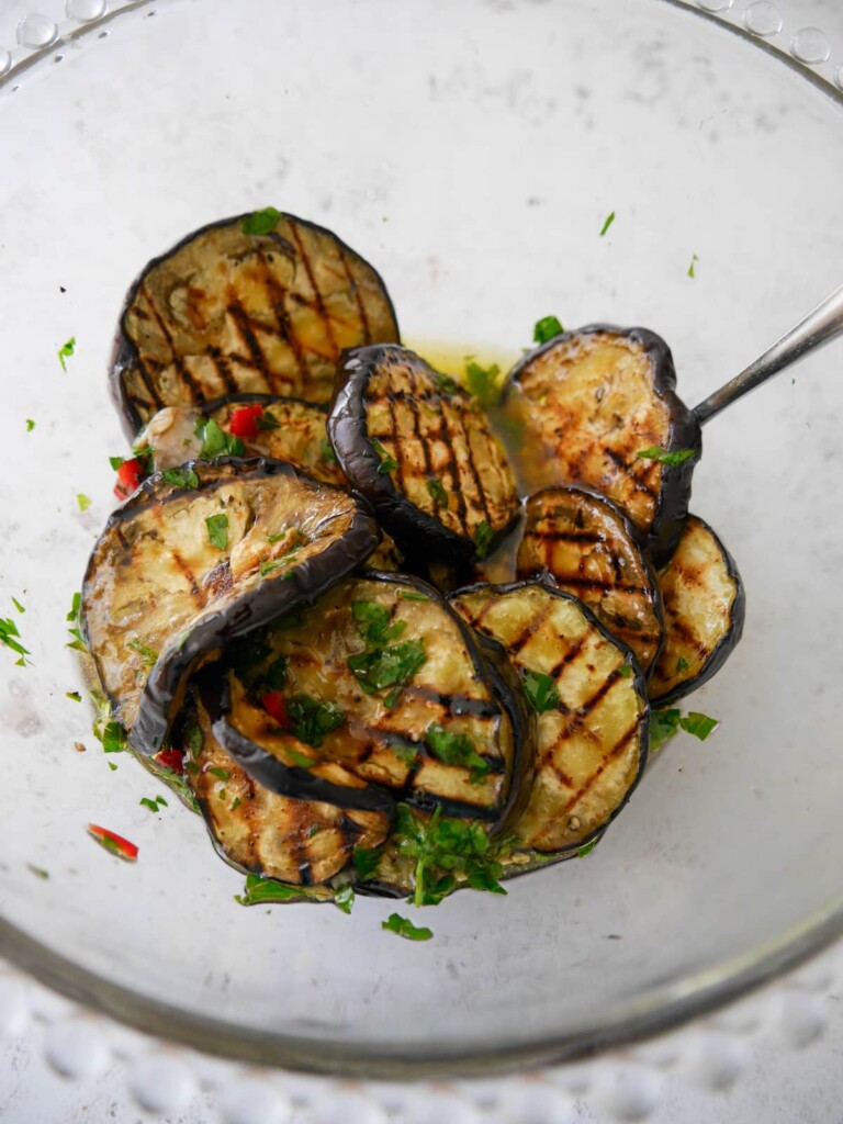 A large bowl of grilled aubergine slices covered in olive oil, chilli, garlic and fresh herbs.
