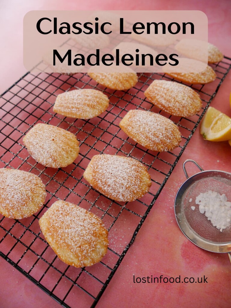 Pinnable image with recipe title and a wire rack of lemon madeleines dusted with a little icing sugar.