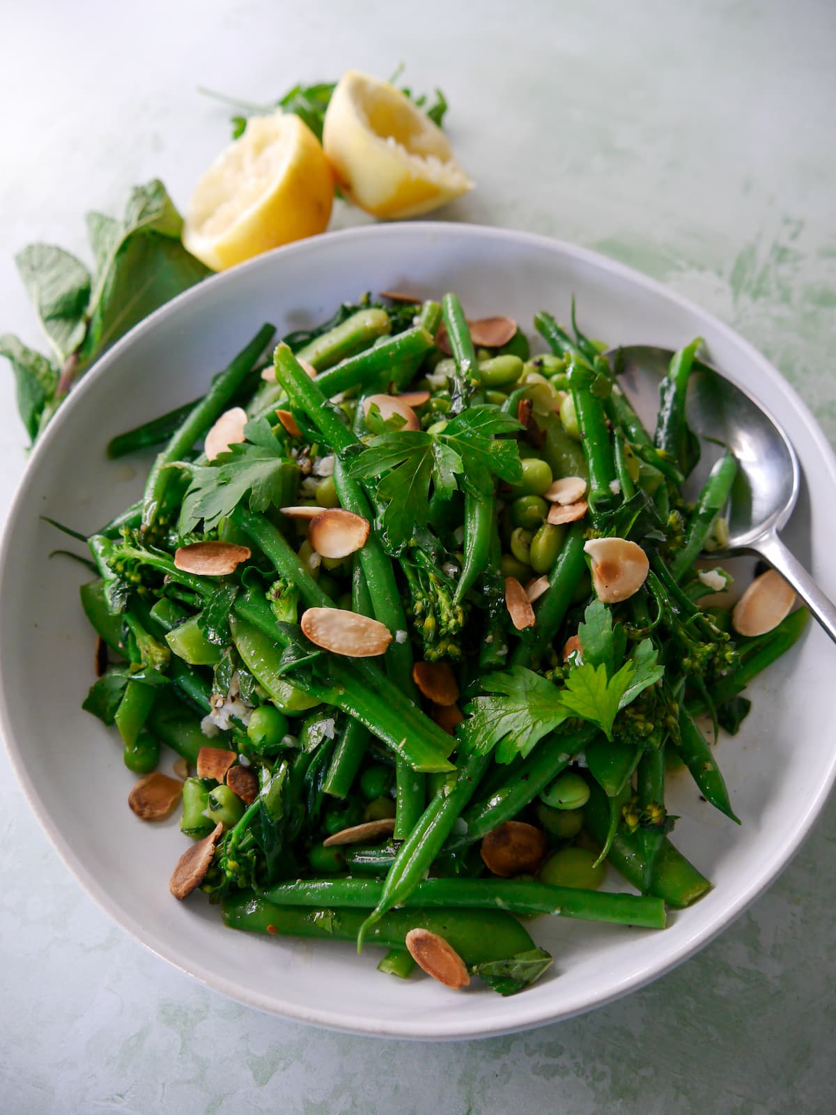 A bowl of mixed green vegetables including tenderstem broccoli, green beans, sugar snap peas, garden peas and edamame beans, coated with a garlic and herb butter with added toasted flaked almonds.