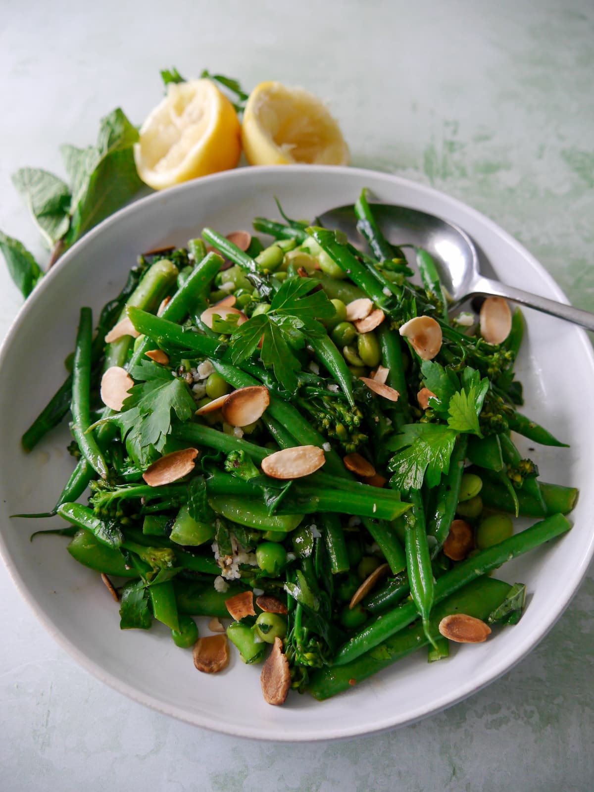 A bowl of mixed green vegetables including tenderstem broccoli, green beans, sugar snap peas, garden peas and edamame beans, coated with a garlic and herb butter with added toasted flaked almonds.