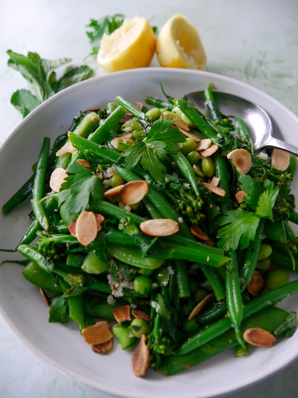 A bowl of mixed green vegetables including tenderstem broccoli, green beans, sugar snap peas, garden peas and edamame beans, coated with a garlic and herb butter with added toasted flaked almonds.