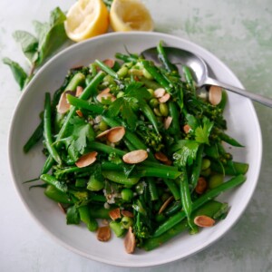 A bowl of mixed green vegetables including tenderstem broccoli, green beans, sugar snap peas, garden peas and edamame beans, coated with a garlic and herb butter with added toasted flaked almonds.