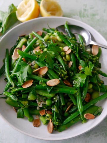 A bowl of mixed green vegetables including tenderstem broccoli, green beans, sugar snap peas, garden peas and edamame beans, coated with a garlic and herb butter with added toasted flaked almonds.