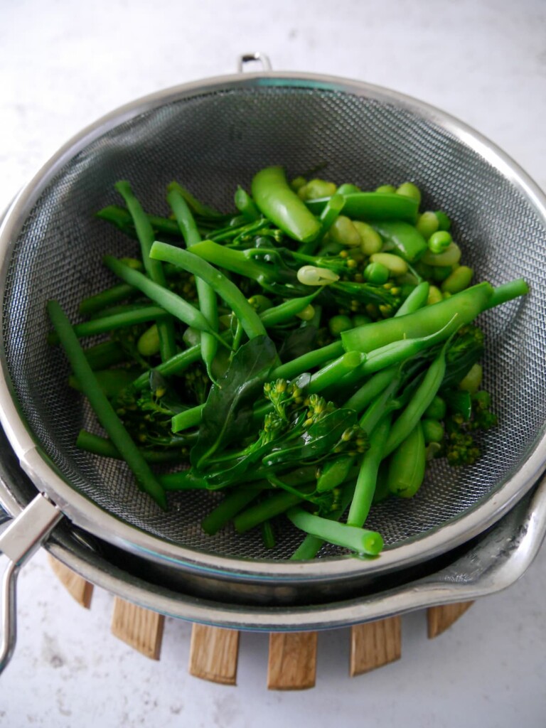 A sieve filled with a mixture of cooked green vegetables draining over a saucepan.