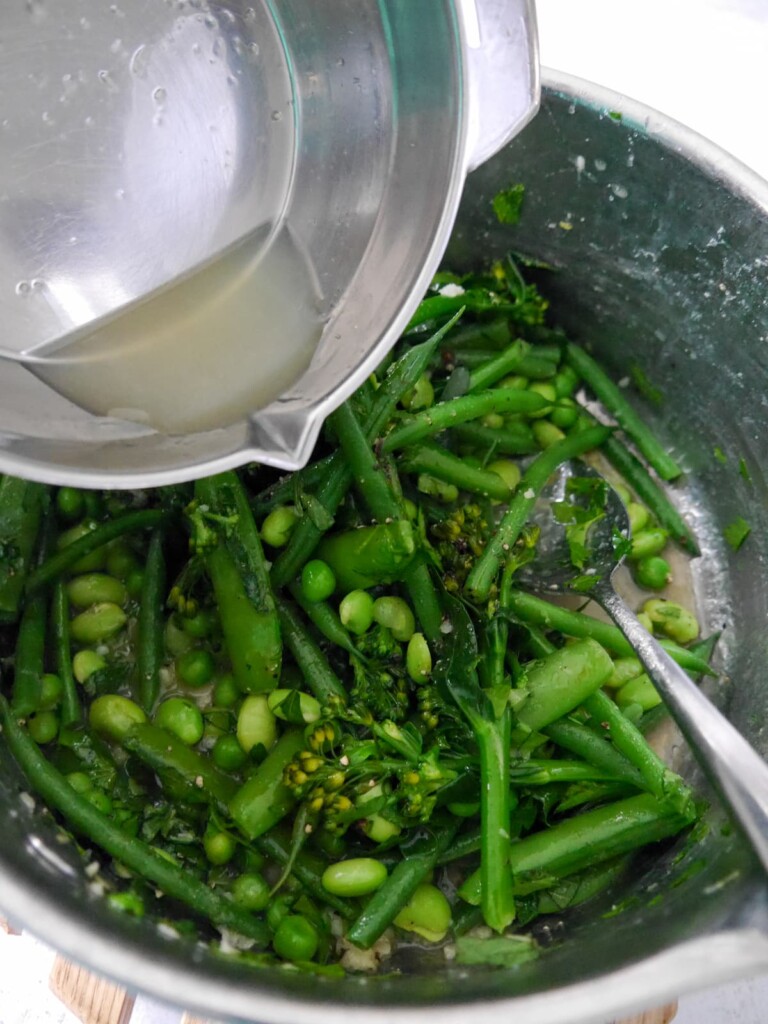 A large saucepan of cooked green vegetables, including tenderstem broccoli, green beans, sugar snap peas, garden peas and edamame beans, coated with a garlic and herb butter with added lemon juice.