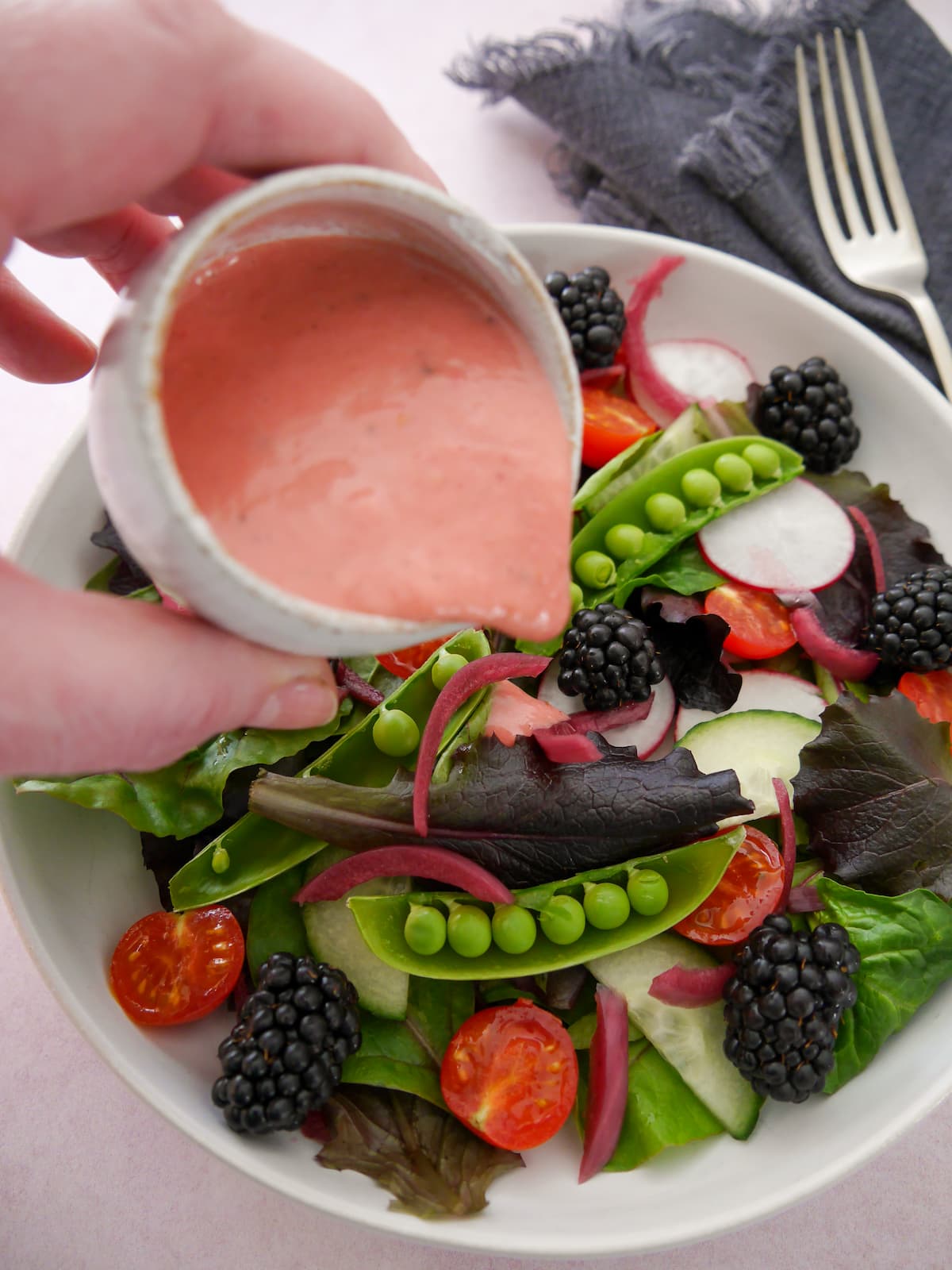 A bowl of mixed salad leaves and vegetables with a small jug of fresh raspberry vinaigrette dressing being poured over.