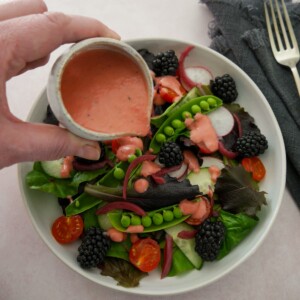 A bowl of mixed salad leaves and vegetables with a small jug of fresh raspberry vinaigrette dressing being poured over.
