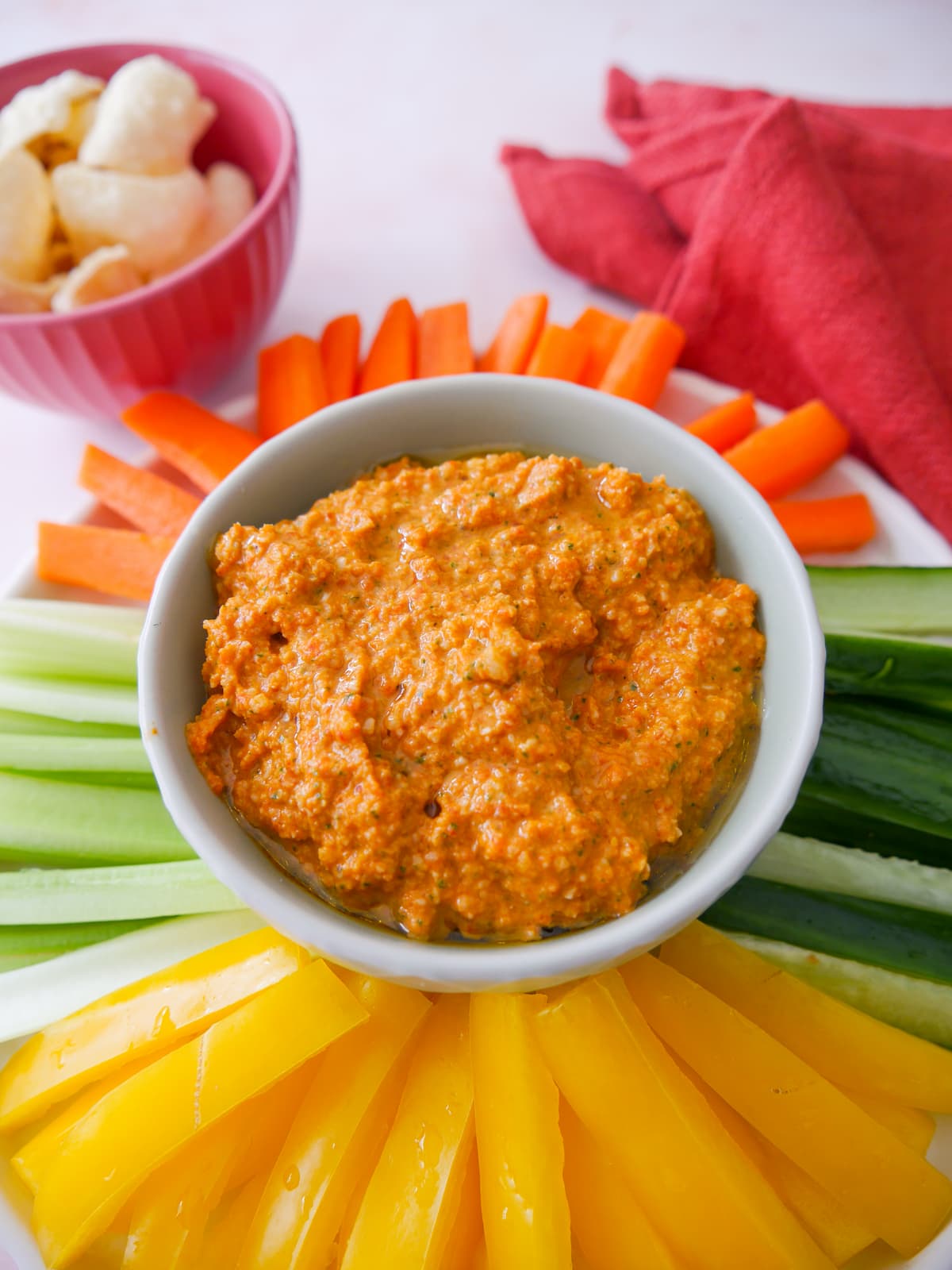 A bowl of roasted red pepper dip served with a variety of fresh vegetable sticks, including bell pepper, cucumber, celery and carrot, with lentil chips served alongside.