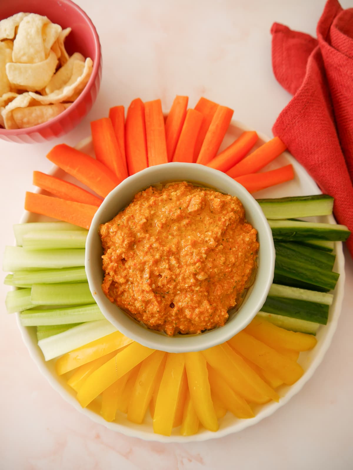 A bowl of roasted red pepper dip served with a variety of fresh vegetable sticks, including bell pepper, cucumber, celery and carrot, with lentil chips served alongside.