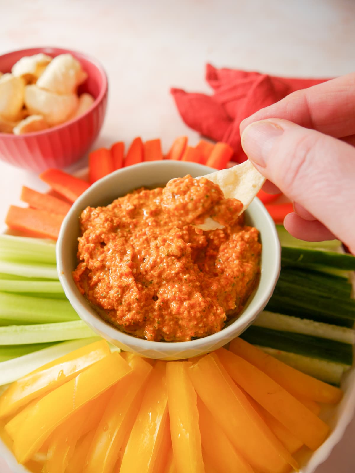 A bowl of roasted red pepper dip served with a variety of fresh vegetable sticks, including bell pepper, cucumber, celery and carrot, with lentil chip being dipped in the bowl.
