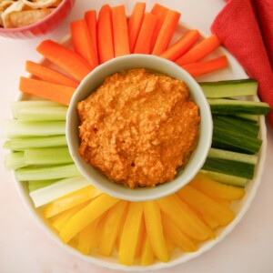 A bowl of roasted red pepper dip served with a variety of fresh vegetable sticks, including bell pepper, cucumber, celery and carrot.