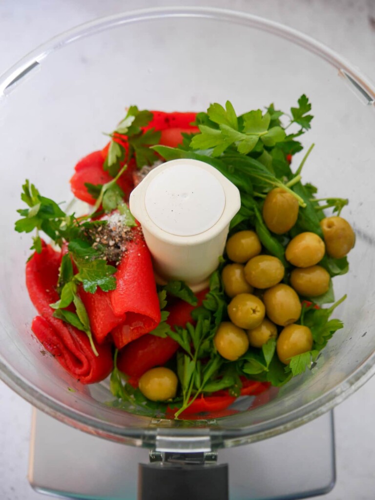 A food processor bowl filled with roasted red peppers, green olives, fresh herbs, olive oil and seasoning.