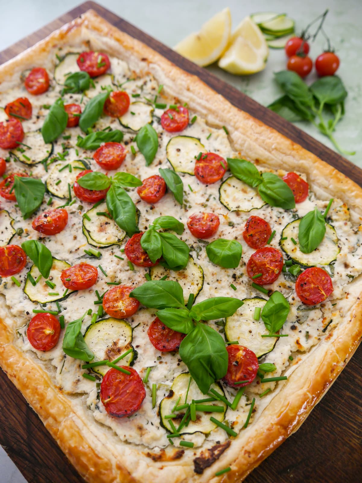 A wooden board topped with a tomato and courgette tart, with ricotta cheese, and garnished with fresh basil and chives.