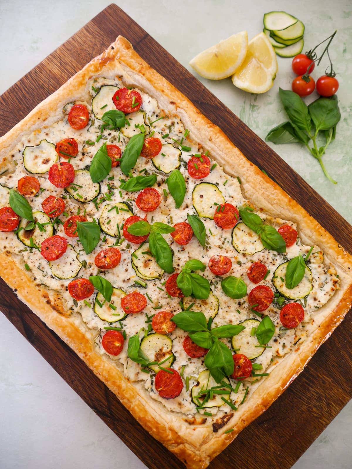 A wooden board topped with a tomato and courgette tart, with ricotta cheese, and garnished with fresh basil and chives.