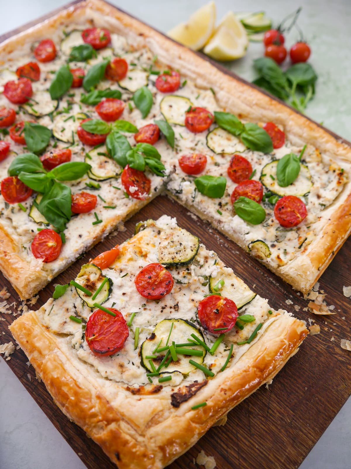 A wooden board topped with a tomato and courgette tart, with ricotta cheese, and garnished with fresh basil and chives, with a slice cut out of the tart.