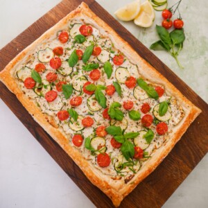 A wooden board topped with a tomato and courgette tart, with ricotta cheese, and garnished with fresh basil and chives.