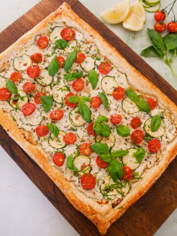 A wooden board topped with a tomato and courgette tart, with ricotta cheese, and garnished with fresh basil and chives.