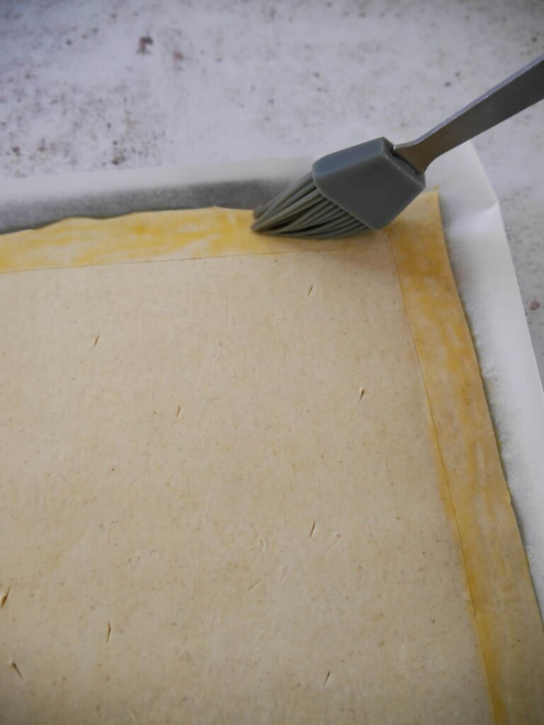 A baking sheet topped with a sheet of puff pastry, with an egg wash being brushed around the edges.