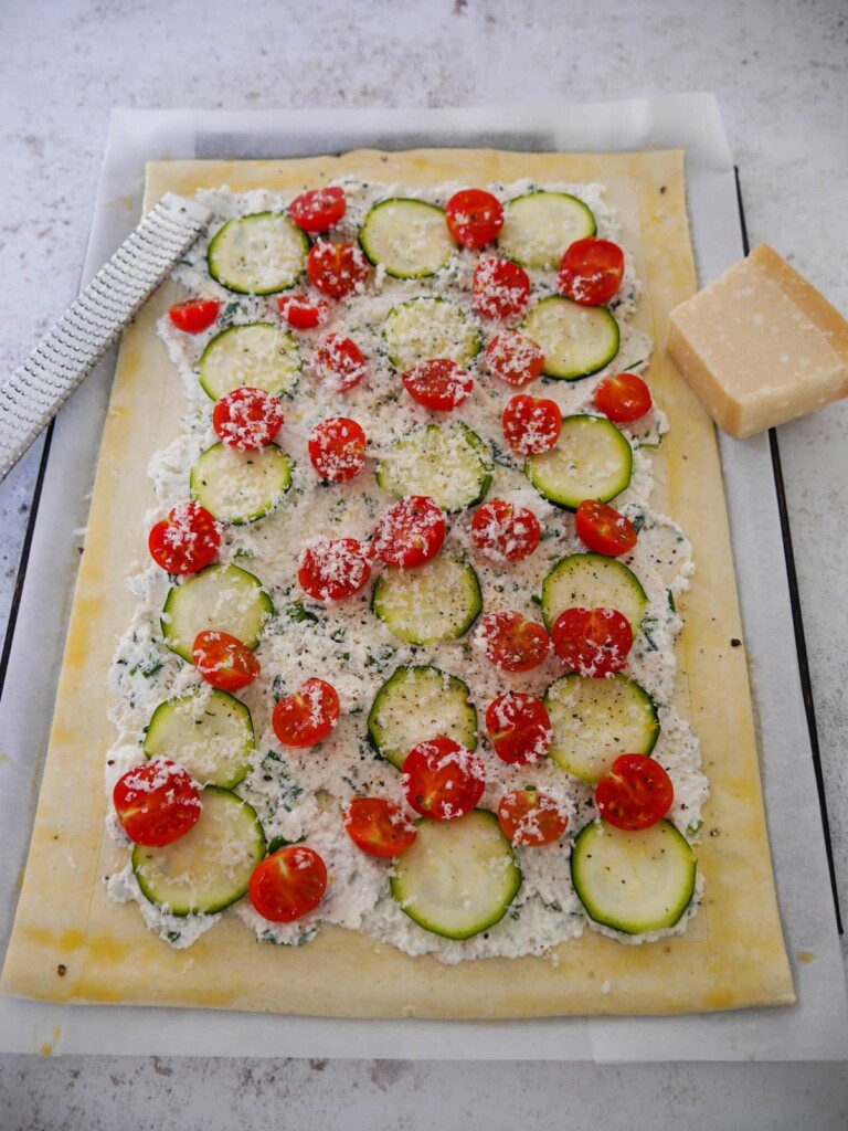 A baking sheet topped with a sheet of puff pastry, topped with a ricotta cheese and herb mixture, thin slices of courgette, cherry tomatoes and finished with a grating of parmesan cheese.
