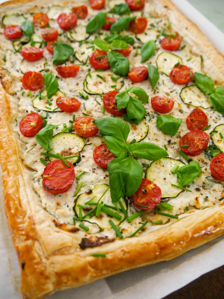 A baking sheet topped with tomato and courgette puff pastry tart, garnished with fresh basil leaves and chives.